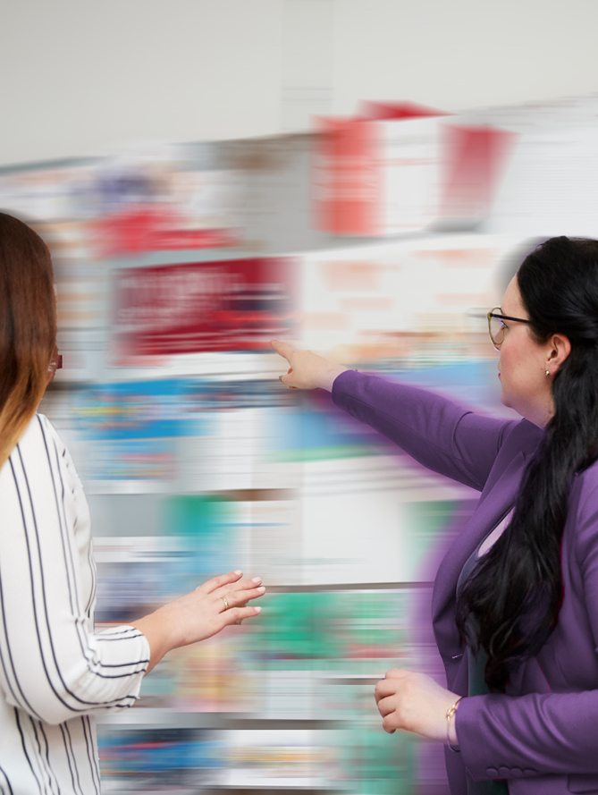 Das Bild zeigt zwei Frauen, die auf eine Wand voller Produkte zeigen. Die Aufnahme ist leicht verschwommen und vermittelt einen Eindruck von Dynamik und Bewegung.